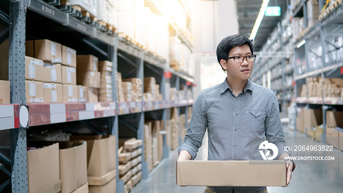 一名年轻的亚洲男子在仓库的一排货架之间搬运纸板箱。购物仓库或