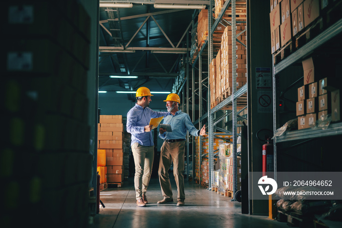 Two successful business partners in formal wear discussing about sales while standing in warehouse.