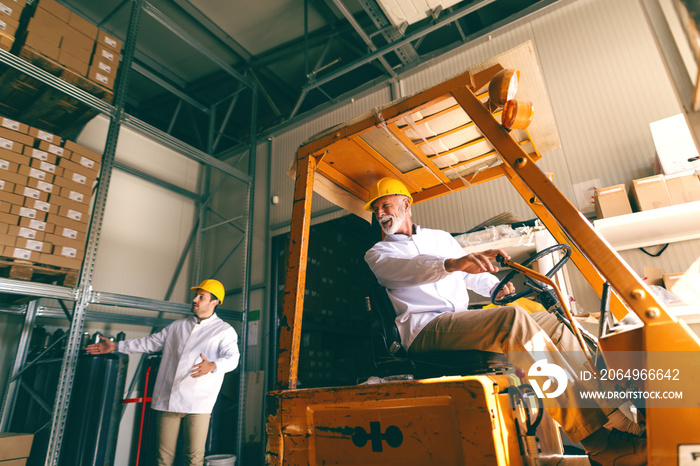 Two workers with helmets on head working in warehouse. Oder one driving forklift while younger one d