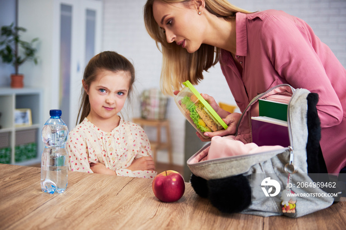 Mother putting lunch box with healthy food in daughters backpack