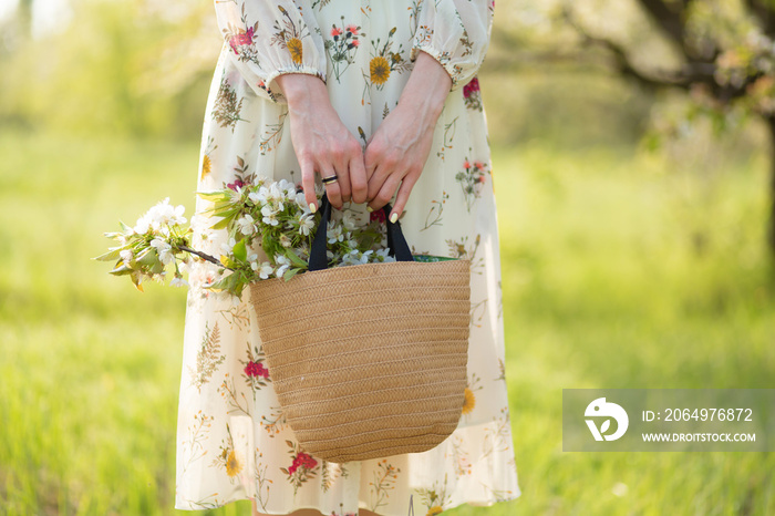 A woman holds in her hands a stylish wicker bag with blooming flowers in green park. Spring romantic