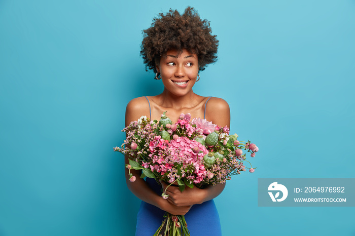 Pretty satisfied dark skinned young woman has Afro hair holds bunch of flowers bites lips and looks 