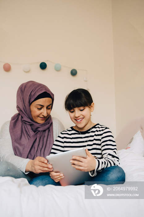 Smiling mother and daughter looking at digital tablet in bed at home