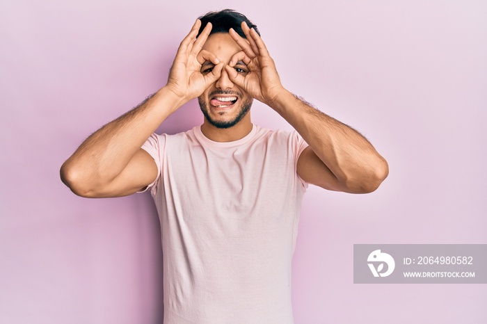 Young arab man wearing casual clothes doing ok gesture like binoculars sticking tongue out, eyes loo