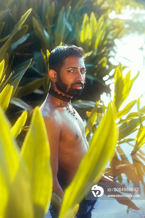 portrait of a dark skinned Indian man by a lake, shirtless, with make up, surrounded by nature