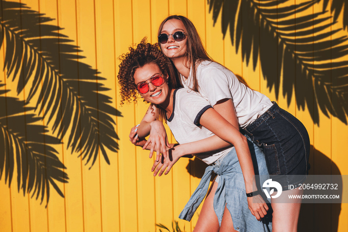 Candid shot of two lovely hipster women in glasses and denim clothes standing under the shadow of pa