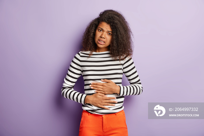 Unhappy Afro American woman touches belly, suffers from menstrual period cramp, has indigestion afte