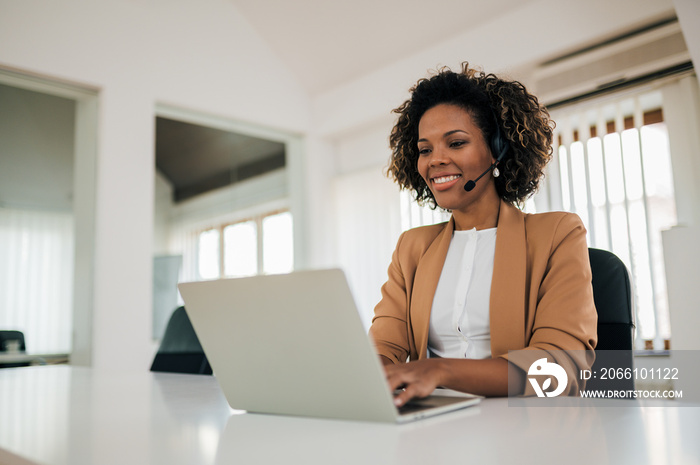 Portrait of a positive mixed race secretary working on laptop, wearing headset.