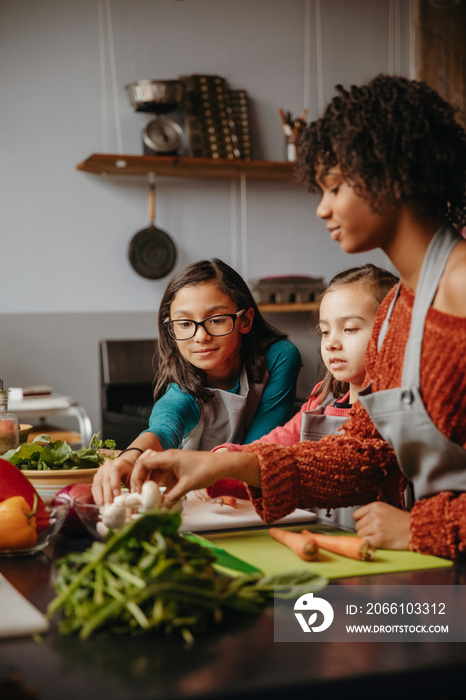 Young woman with cute students preparing vegetables on table against wall in cooking class