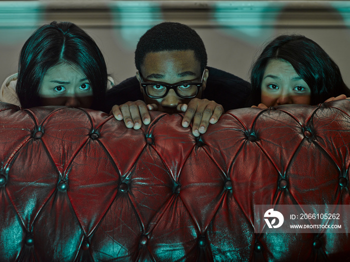 Three young adult friends hiding behind sofa