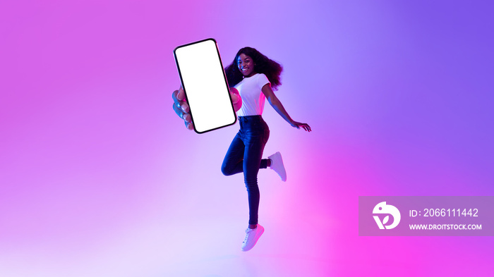 充满欢乐的年轻黑人女性拿着智能手机跳跃，在霓虹灯下显示空屏幕，