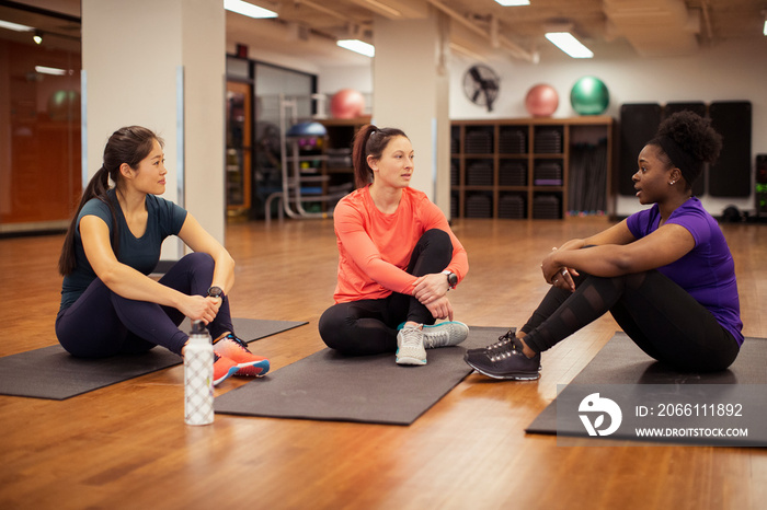 Full length of confident female athletes talking while sitting on exercise mats in gym