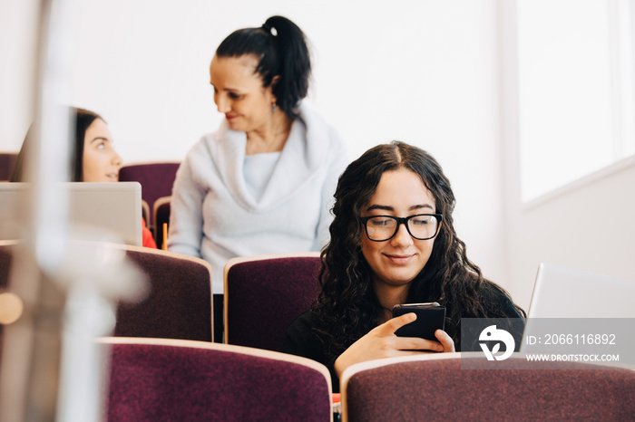 Female student using mobile phone with teacher in background at university