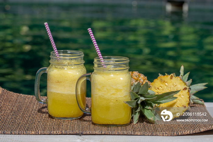 Homemade mango and pineapple smoothie made with coconut milk in two glass mug near swimming pool . I