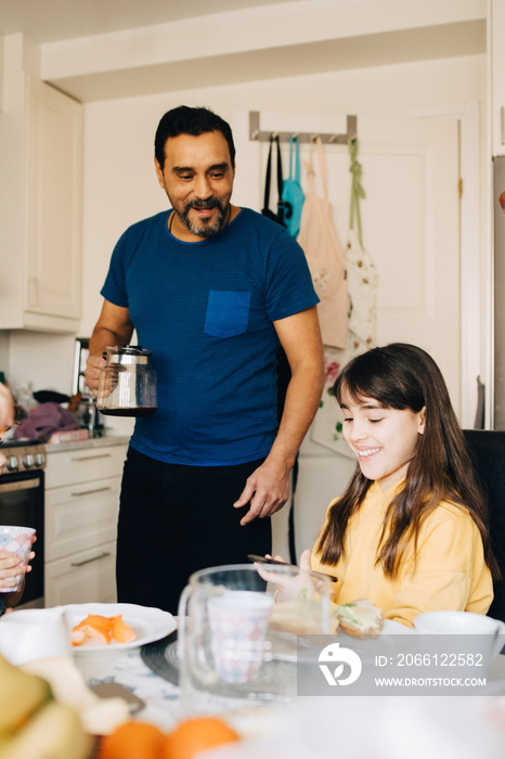 Father with coffee pot looking at daughter using smart phone during breakfast