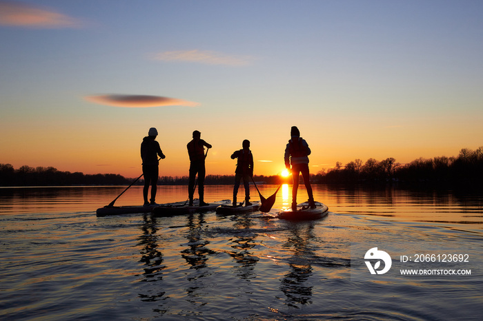 Silhouettes of the people on stand up paddle board at dusk on a flat quiet winter river with beautif