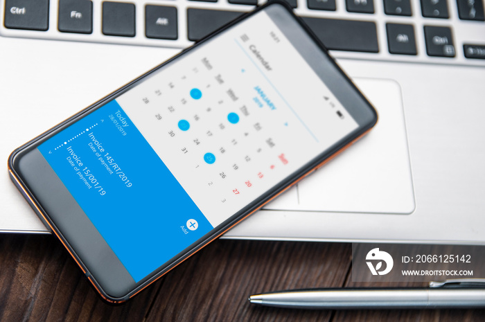 Smartphone witch notch lies on the desk with calendar application that remind about invoices to pay.