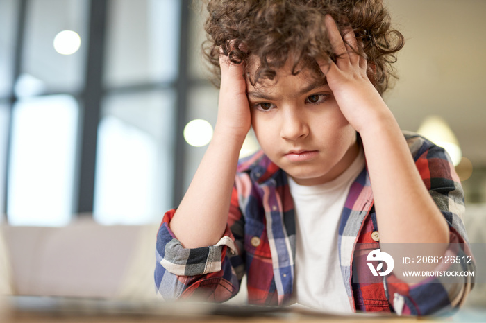 Too much to learn. Portrait of tired latin boy holding his head, looking sad while sitting at the de