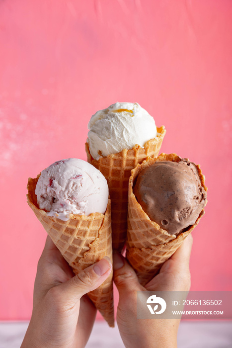 hands holding strawberry, vanilla, chocolate ice cream with waffle cone on pink backgrounds