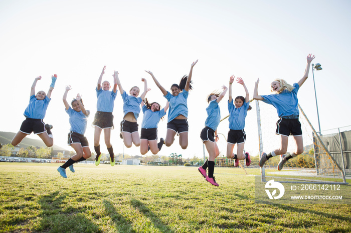 Happy girls in soccer uniforms with arms raised jumping on grassy field against sky