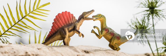 selective focus of toy dinosaurs roaring on sand dune with tropical leaves, panoramic shot