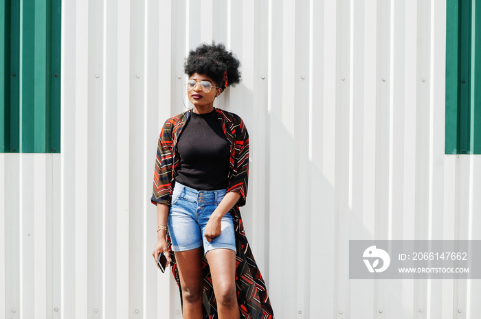 African woman with afro hair, in jeans shorts and eyeglasses posed against white steel wall with mob