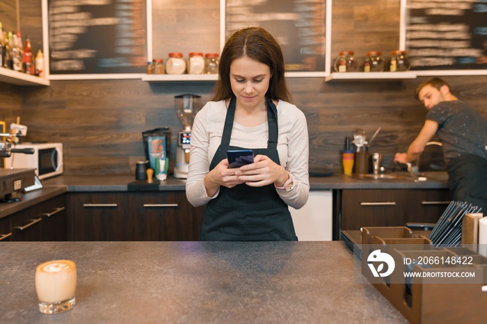 Young woman in apron coffee shop worker at bar counter, taking order on smartphone