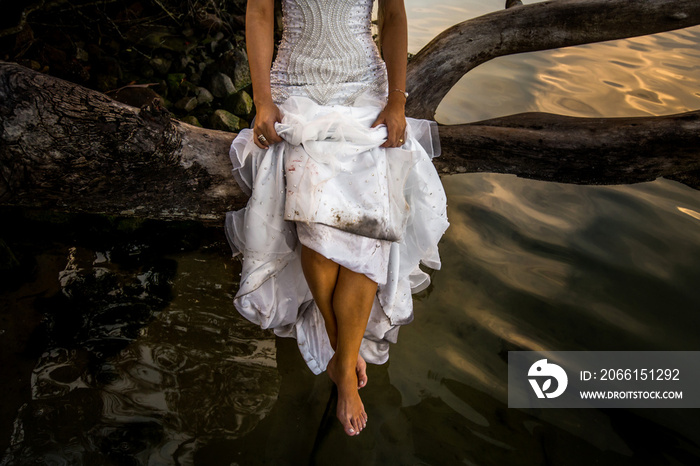 Uma mulher, com seu vestido de noiva, sentada em um galho de árvore, por cima da água escura de uma 