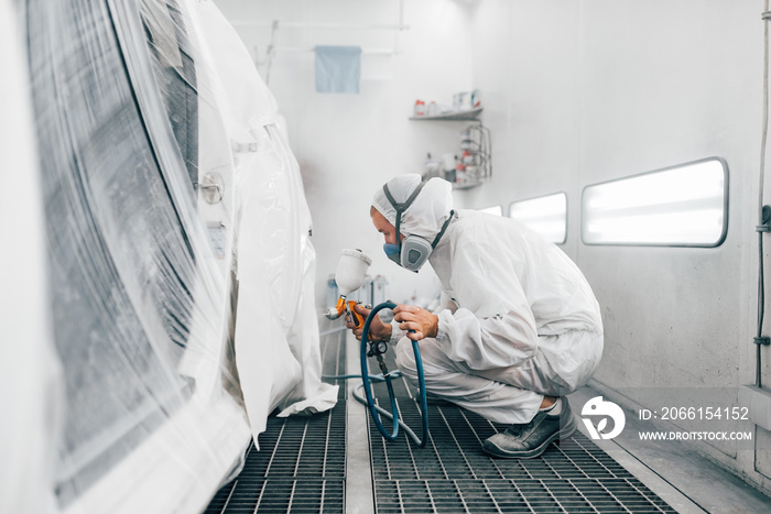 Worker in protective uniform painting a car in a paint booth.