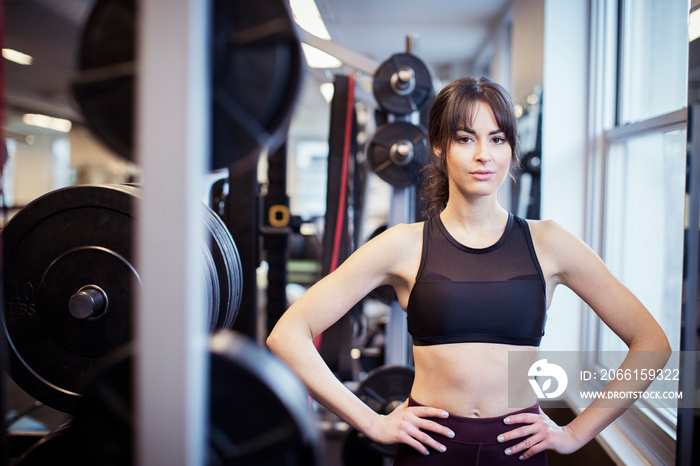 Portrait of confident female athlete with arms akimbo standing by exercise equipment in gym