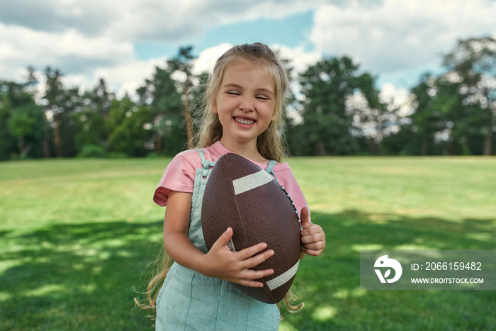 Portrait of happy little girl holding an oval brown leather rugby ball and smiling while playing wit