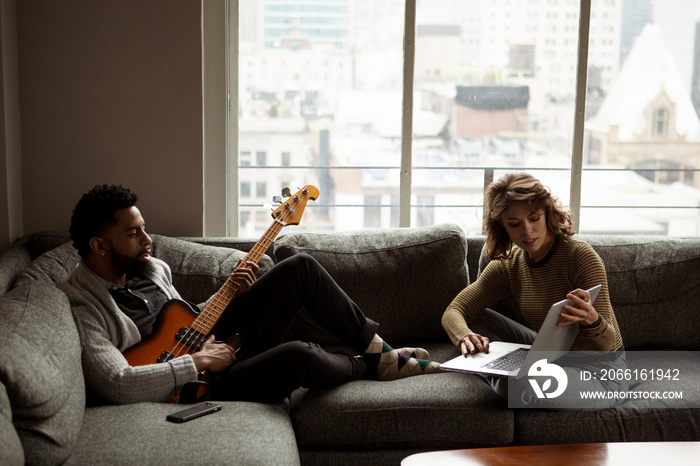 Businesswoman showing laptop computer to businessman playing guitar while relaxing on sofa against w