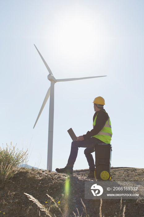 Male engineer with laptop working below sunny wind turbine