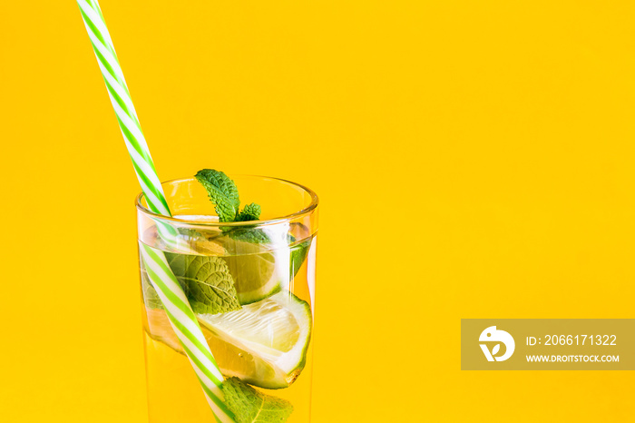 Fresh summer lemonade with water, lime and mint in a glass tumbler with a straw on a yellow backgrou