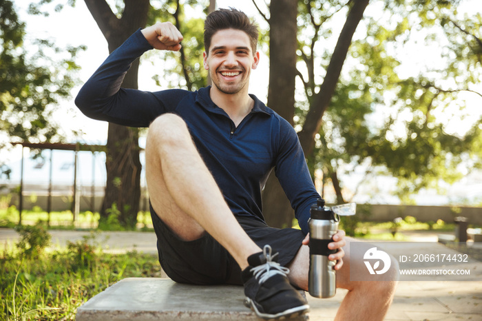 Portrait of strong athletic man sitting on bench and showing his bicep while doing workout in sunny 
