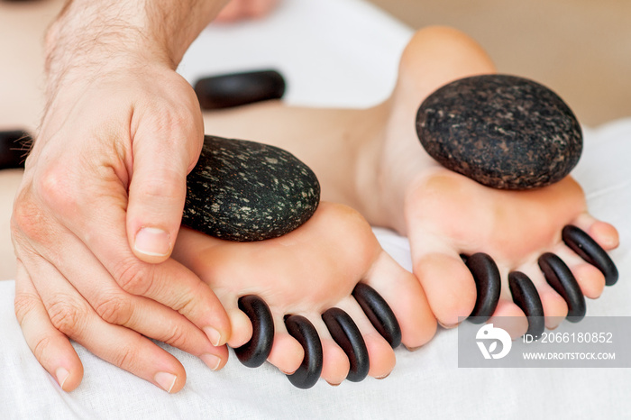 Hands of masseur putting hot little black stones between female toes for feet massage