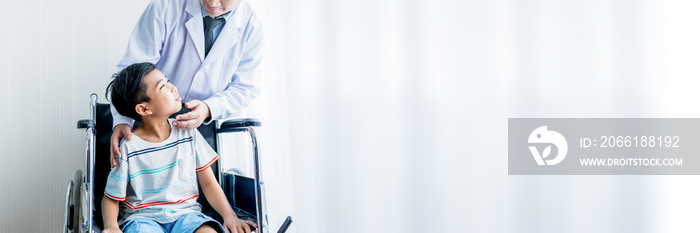 Male doctor standing, caring and cheering for a boy patient sitting in a wheelchair by the window. W