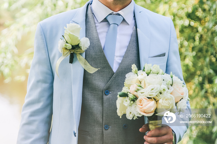 Groom in blue suit is standing outdoors. Handsome man is holding the wedding bouquet.