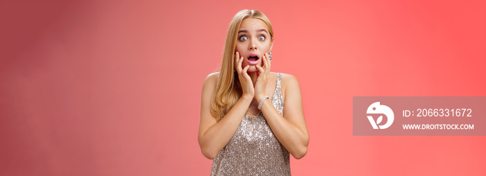 Shocked concerned stunned young european blond woman in silver dres drop jaw widen eyes stunned speechless gasping open mouth touch face amazed, look troubled perplexed, red background