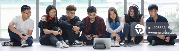 Group of seven young male and female teenagers in casual clothes sitting on the floor and looking at the tablet screen with attractive smiling. College student playing games together with controllers