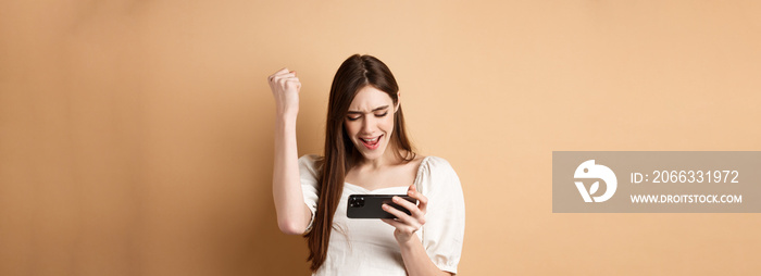 Euphoric happy winner playing mobile video game, shouting yes with joy and making fist pump, winning online, looking at smartphone screen, beige background