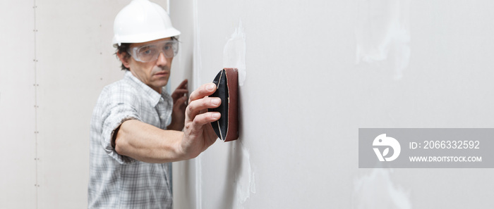 man drywall worker or plasterer sanding and smoothing a plasterboard walls with stucco using a sandpaper holder. Wearing white hardhat and safety glasses. Panoramic image with copy space