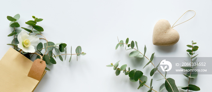 Craft bag with wintertime eucalyptus and white Helleborus flower. Panoramic banner. Brown paper tag, copy-space. Winter flat lay with evergreen twigs and winter rose flower on off white background