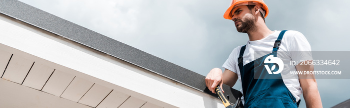 panoramic shot of repairman in helmet and uniform holding hammer and standing against blue sky with clouds