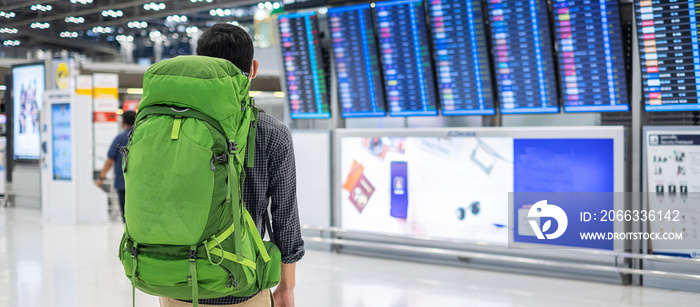 Young man traveler with backpack checking flight time, Asian passenger with hat looking to information board in international airport terminal. Travel, vacation and world tourism day concept