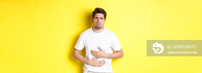 Man having stomach ache, grimacing from pain and touching belly, standing over yellow background