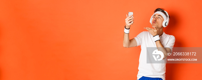 Portrait of attractive middle-aged man in gym uniform, wearing headphones, sending air kiss at mobile phone camera, taking selfie or having video call, standing over orange background