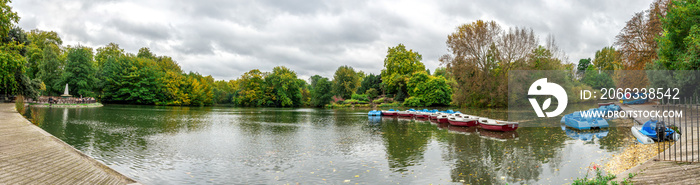 Panoramic view of a boating lake with paddle boats parked and park cafe outdoor tables, Battersea Park, London