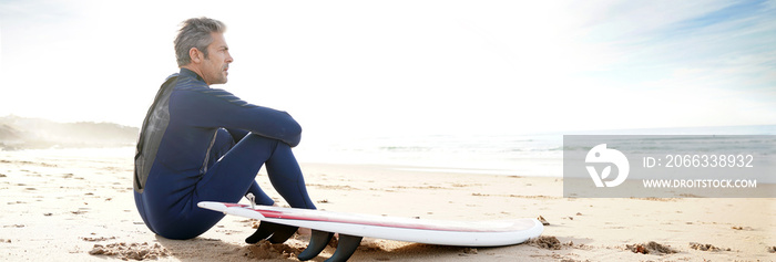 Surfer sitting on sandy beach, next to surfboard