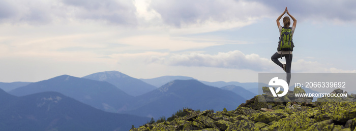 Back view of young slim tourist girl with backpack standing on one leg in yoga pose on rocky top on bright blue morning sky and foggy mountains background. Tourism, traveling and climbing concept.